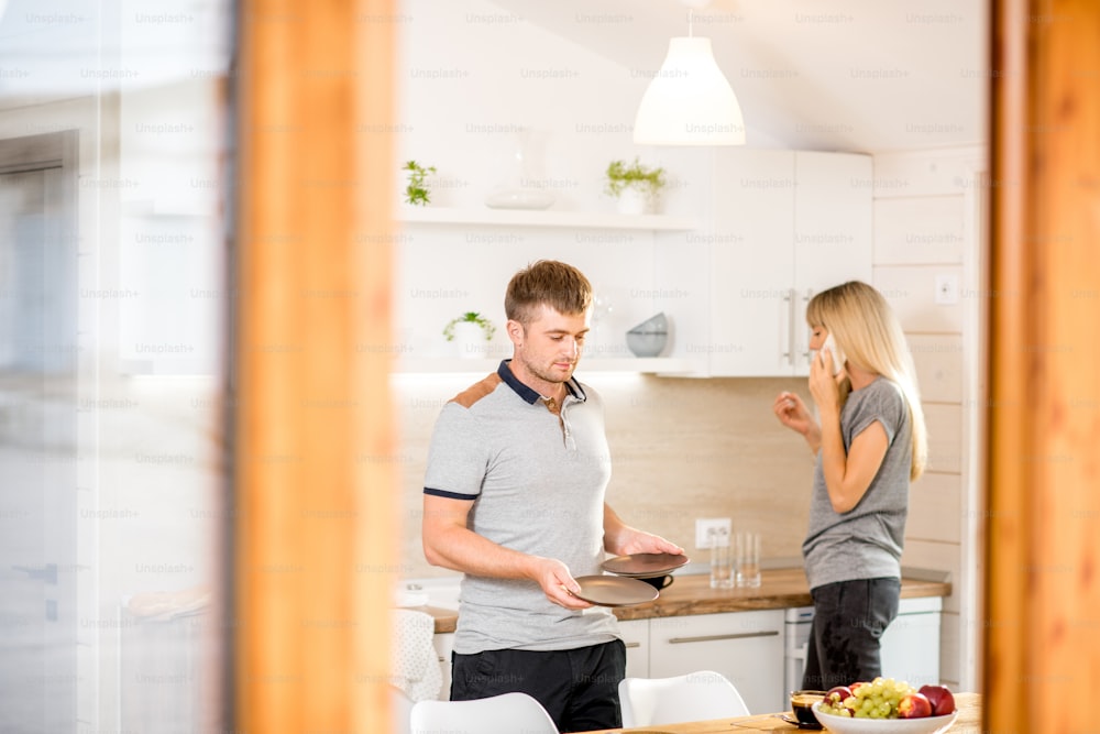 Giovane coppia che serve piatti e parla al telefono che si prepara per la cena nella cucina della moderna casa di campagna