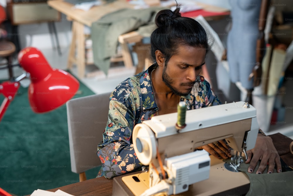 Colpo ad angolo alto di giovane designer di vestiti di etnia indiana seduto al tavolo usando la macchina da cucire per fare il vestito per la nuova collezione