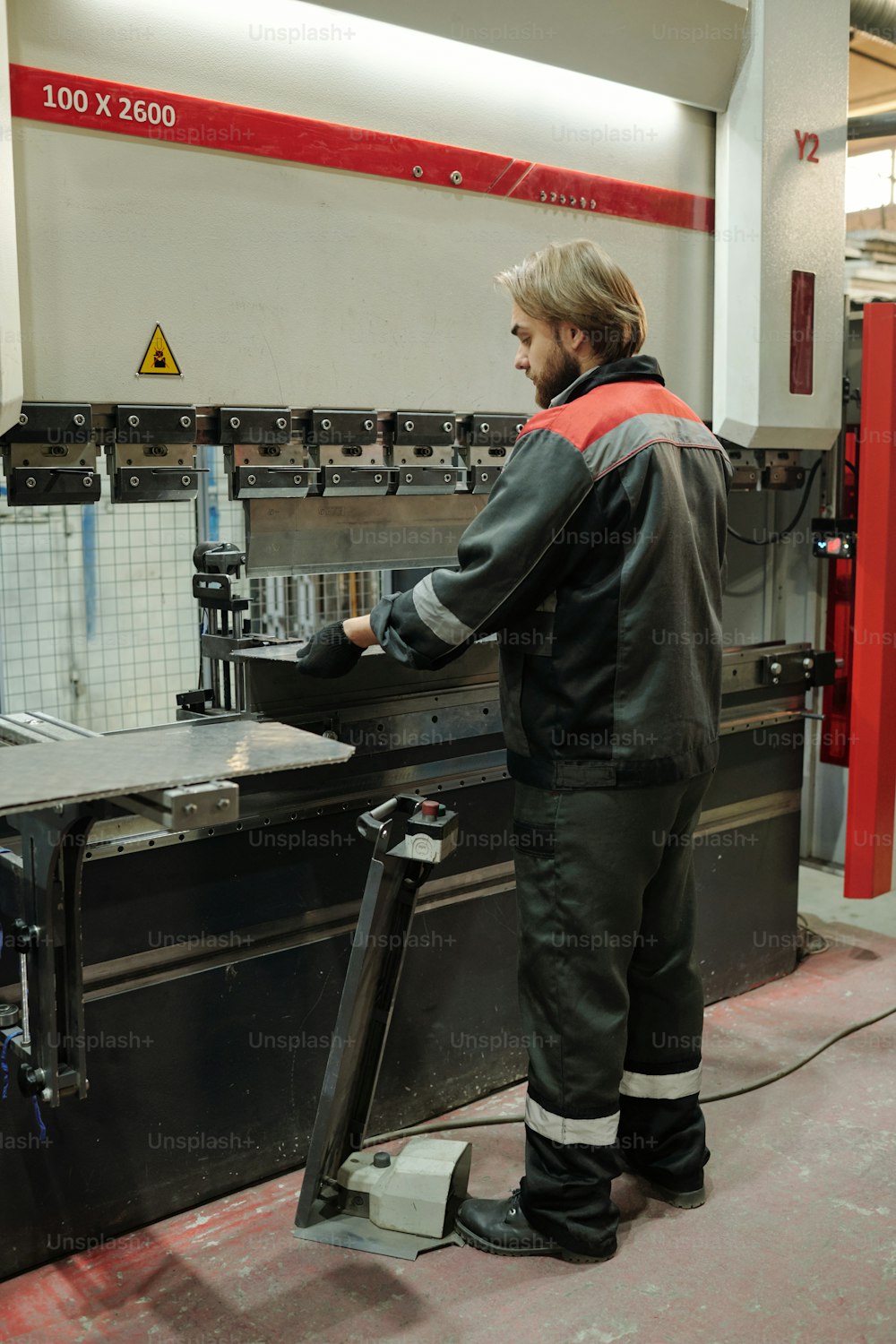 作業場で巨大な産業機械の前で作業しながら、メカニズムのペダルを踏む作業服を着た若い男性の側面図