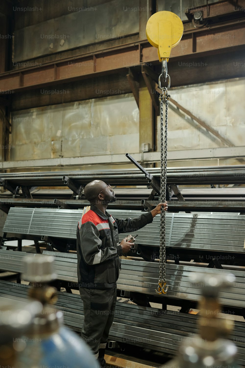 Jovem negro puxando um monte de correntes metálicas enquanto está em pé na frente de pilhas de materiais para a construção de máquinas industriais