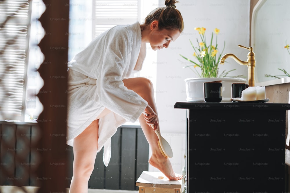 Young woman in white bathrobe doing body massage with dry wooden brush with natural bristles in bathroom at home