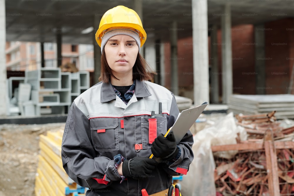 Young serious female engineer in hardhat and uniform holding document and looking at camera while standing against unfinished building