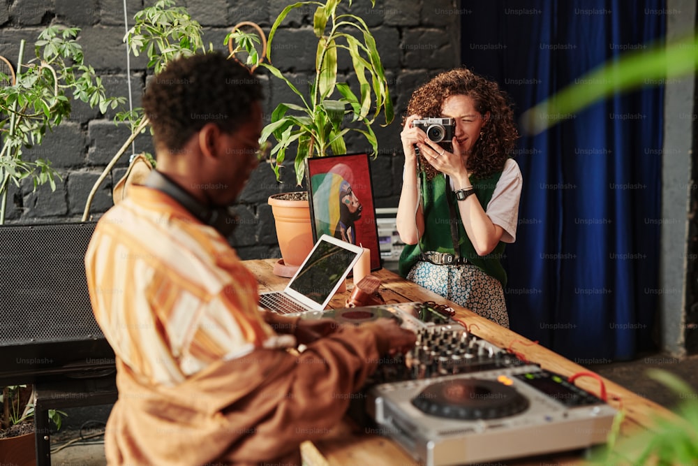 Chica con cámara de fotos tomando foto de su novio afroamericano de pie junto a la mesa de madera con dj set y creando nueva música