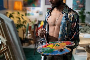 Jovem atraente vestindo camisa desabotoada segurando palete de madeira com tintas a óleo e pincel criando novas obras de arte na tela