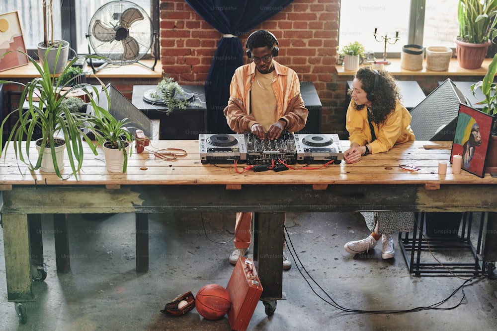 Joven pareja interracial con auriculares y ropa informal creando nueva música mientras un hombre negro ajusta los sonidos en el controlador de dj