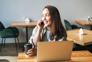Femme d’affaires brune souriante adulte de quarante ans aux cheveux longs en chemise élégante travaillant sur un ordinateur portable à l’aide d’un téléphone portable dans un café
