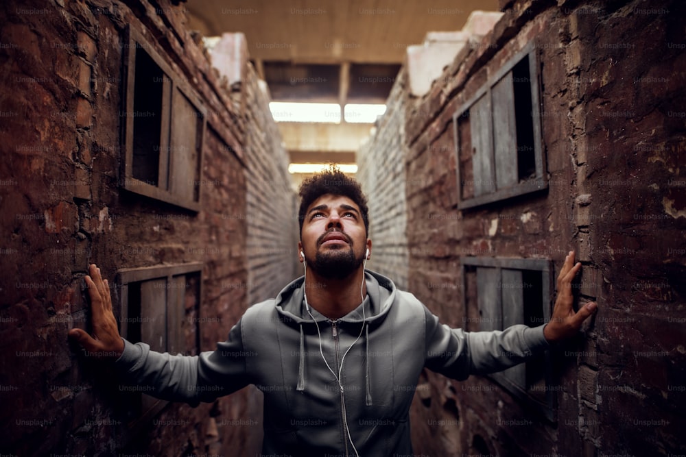Retrato de um jovem atlético atraente e motivado focado com fones de ouvido em pé dentro do lugar abandonado no meio de duas paredes e olhando para cima.