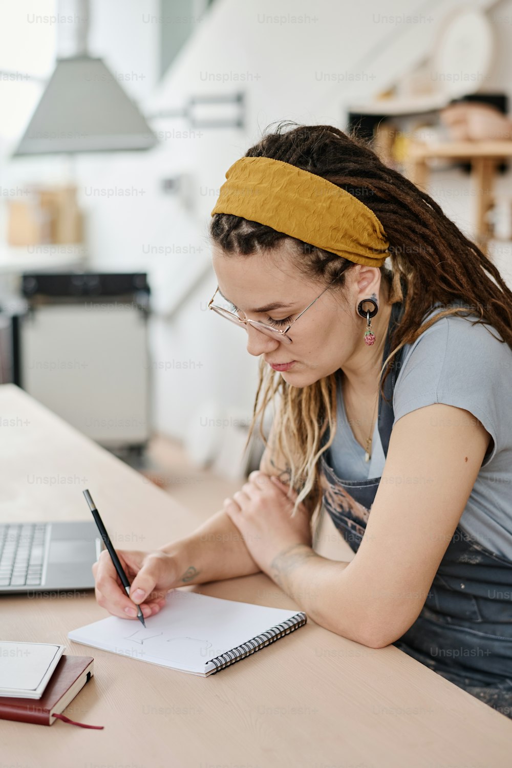 Giovane femmina creativa che fa appunti o schizzo di nuovo prodotto in terracotta nel blocco note mentre si siede sul posto di lavoro in studio