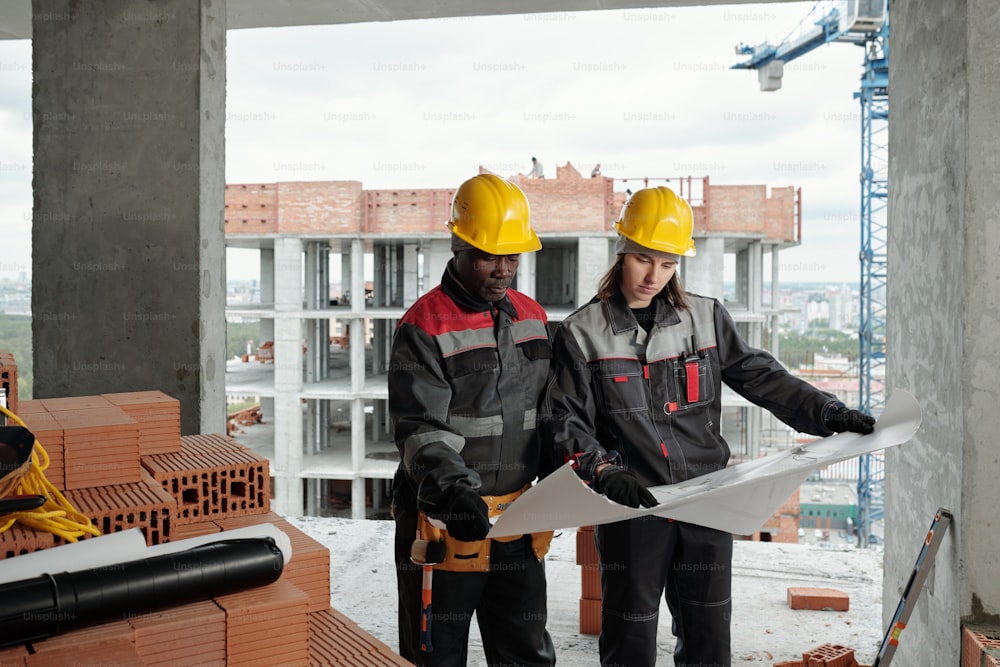 Dois engenheiros interculturais ou construtores em roupas de trabalho e capacetes discutindo plano arquitetônico ou esboço de construção em planta