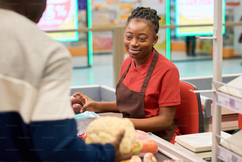 Happy young black woman in uniform sitting by counter and scanning food products while serving male customer in supermarket