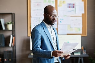 African businessman in eyeglasses examining business documents while standing at office