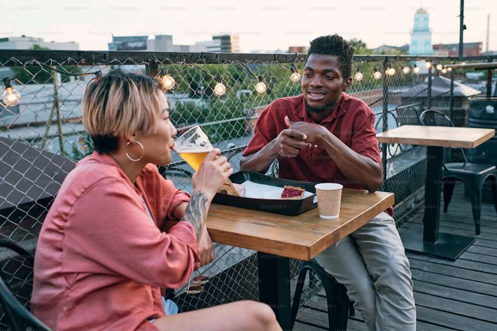Jeune couple interculturel ayant de la restauration rapide et des boissons tout en se relaxant à table dans un café en plein air
