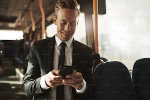 スーツを着た笑顔の若いビジネスマンが朝の通勤中にバスに立ってテキストメッセージを読んでいます