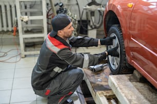Joven reparador en ropa de trabajo colocando rueda nueva en el automóvil mientras trabaja en el garaje del servicio de mantenimiento