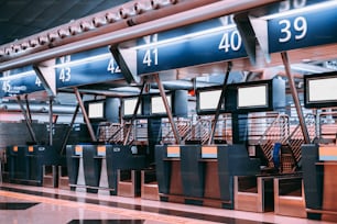 近代的な空港のチェックインエリアの内部:手荷物処理ベルトコンベアシステムを備えた手荷物受入ターミナル、複数の空白の白い情報LCDスクリーンテンプレート、インデックス付きチェックインデスク