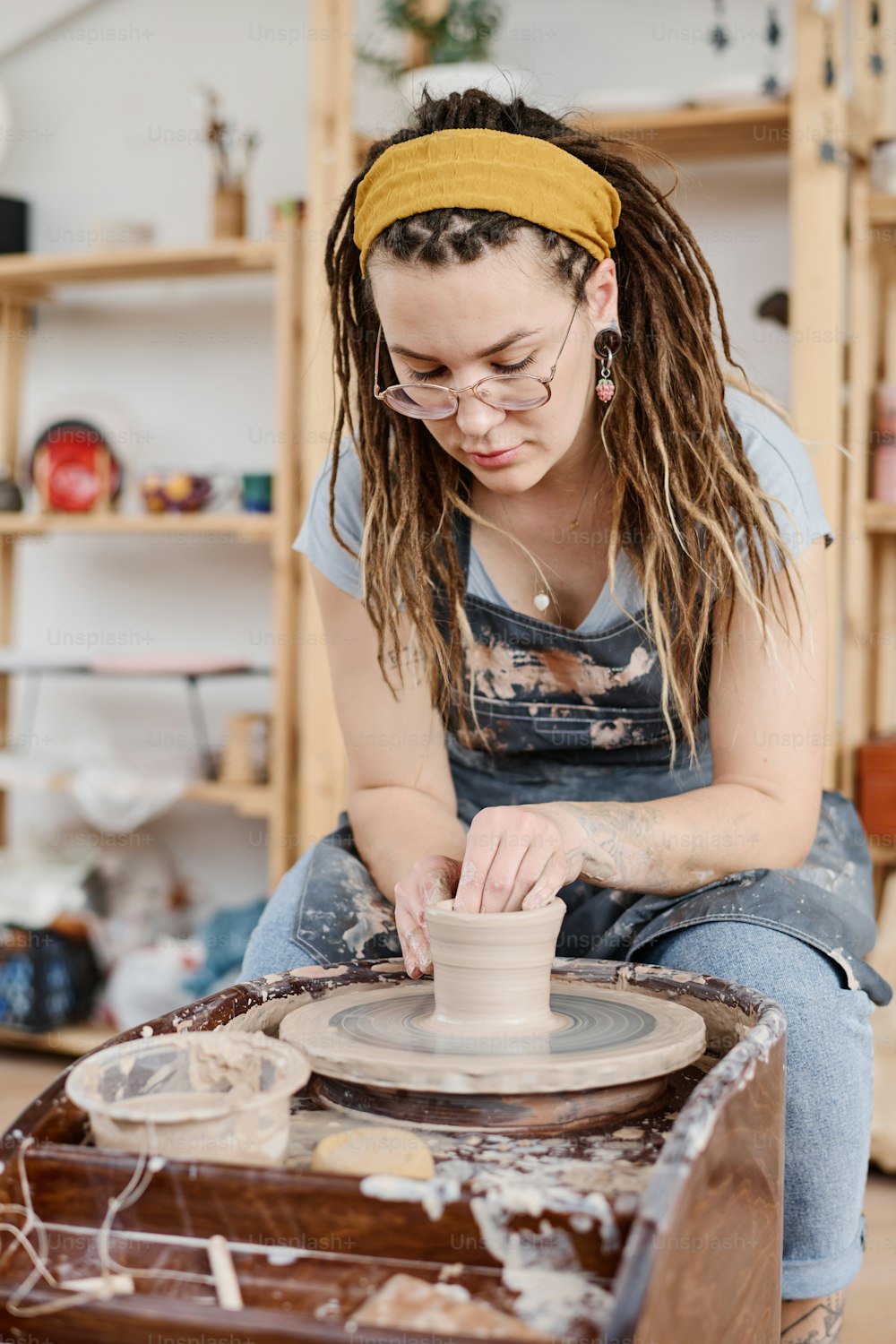 Junge kreative Frau in Arbeitskleidung sitzt vor rotierender Töpferscheibe in der Werkstatt und stellt neue Steingutartikel zum Verkauf her