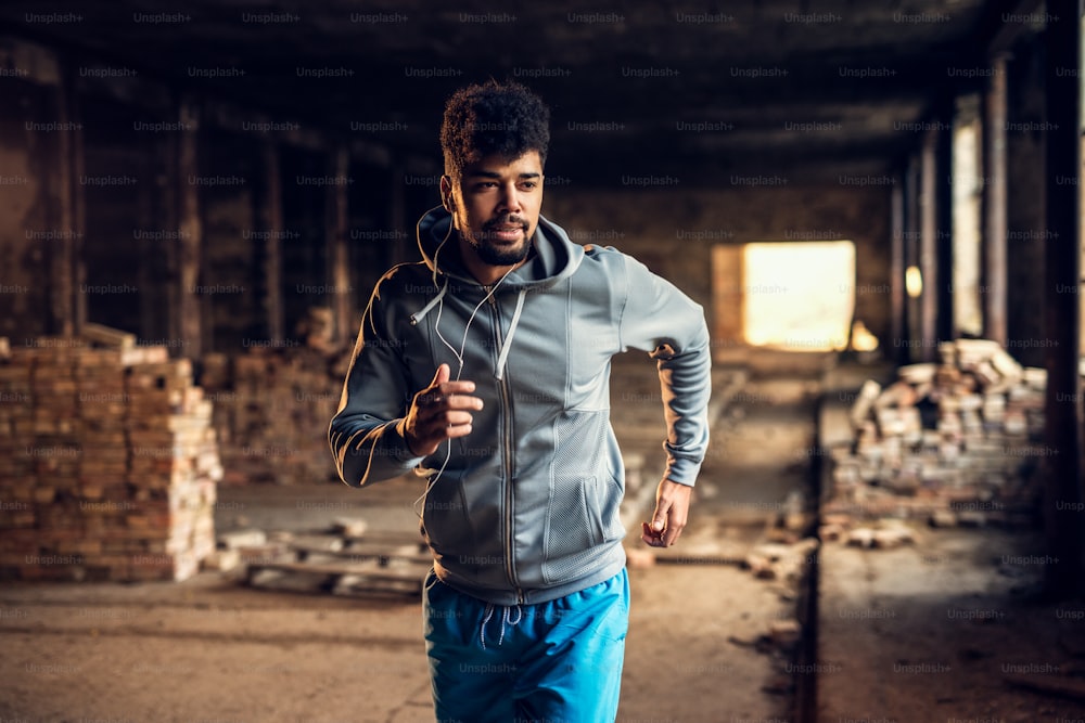 Retrato de um jovem atlético atraente e motivado e motivado com fones de ouvido correndo dentro do lugar abandonado.