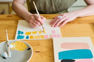 Mãos da mulher nova com pincel fazendo amostras de cor para sua nova pintura enquanto sentado ao lado da mesa de madeira na oficina
