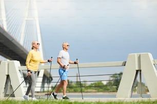 Mature couple with trekking sticks walking along bridge by riverside in the morning