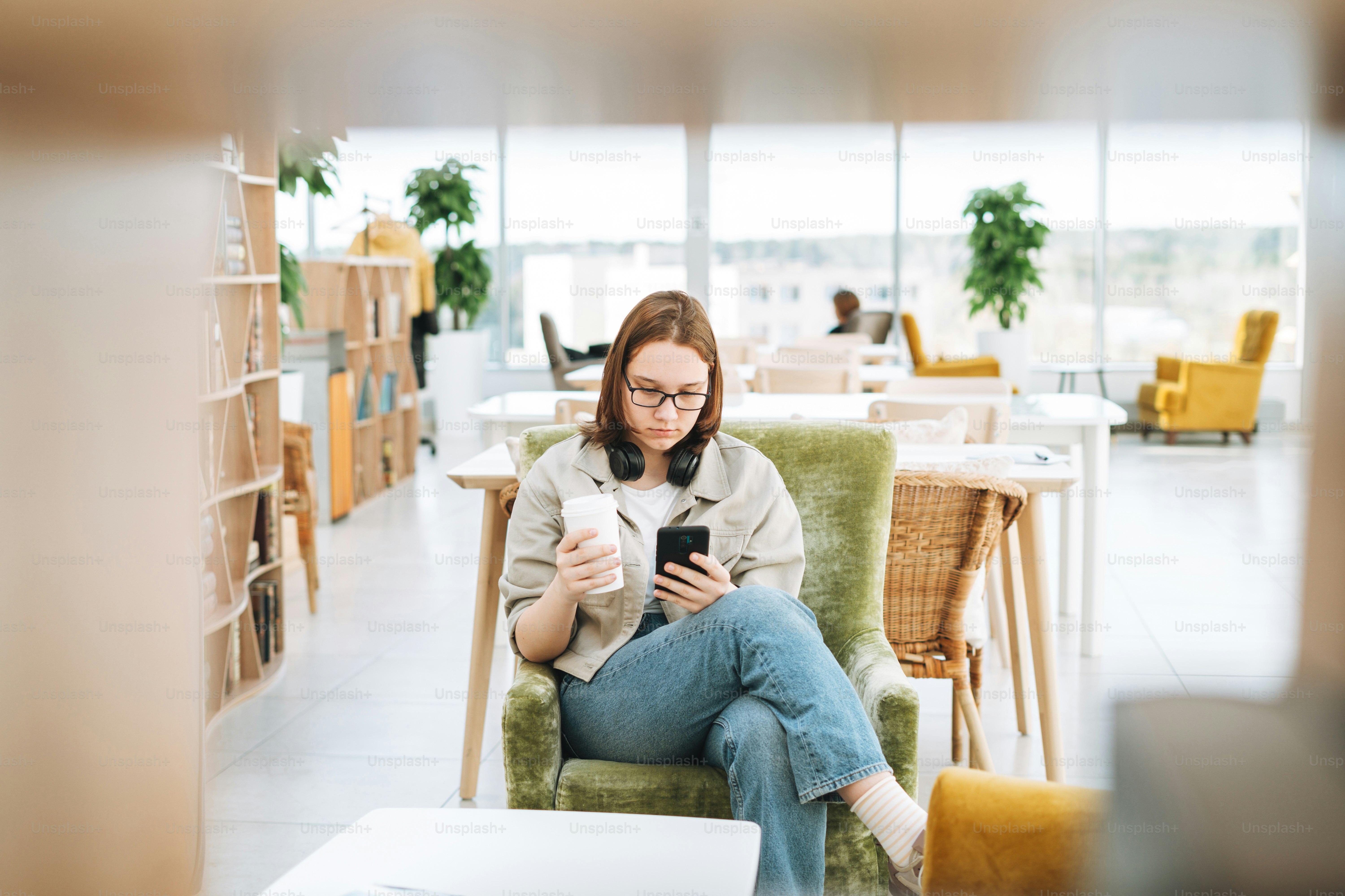 Young brunette teenager girl college or high school student in glasses using mobile phone working at green modern library, public place, green open space office
