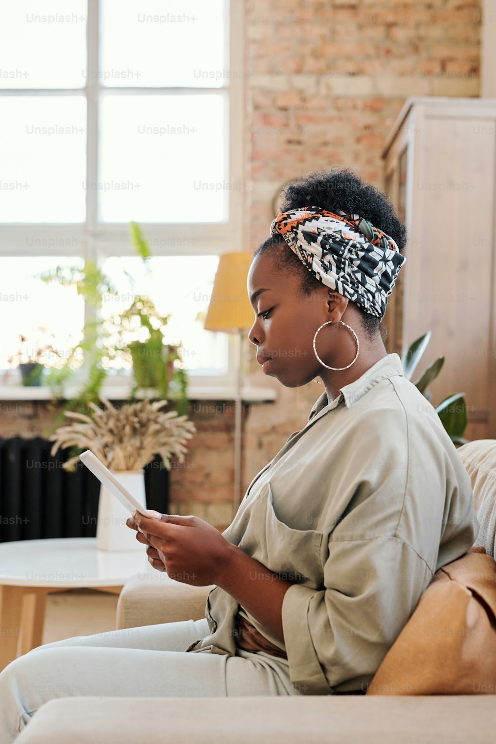 Vista laterale di una giovane donna nera seria in foulard seduta in poltrona e che naviga in internet mentre utilizza il tablet