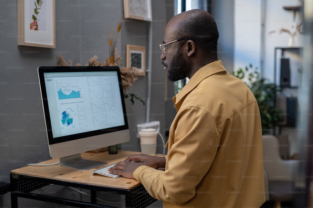 Hombre de negocios afroamericano moderno sentado junto al escritorio frente al monitor de la computadora y mirando el diagrama financiero en la pantalla