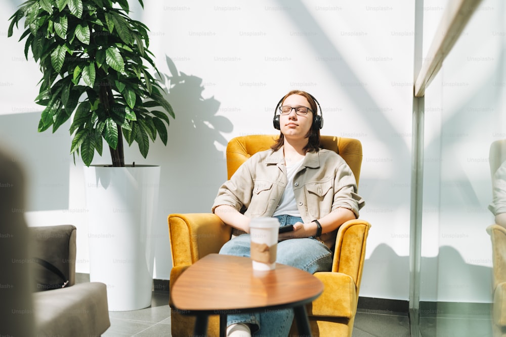 Young brunette teenager girl college or high school student in glasses using mobile phone, listen music in headphones in yellow chair at public place