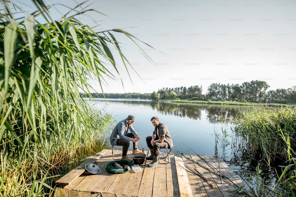 Two fishermen relaxing during the picnic on the beautiful wooden pier with green reed on the lake in the morning