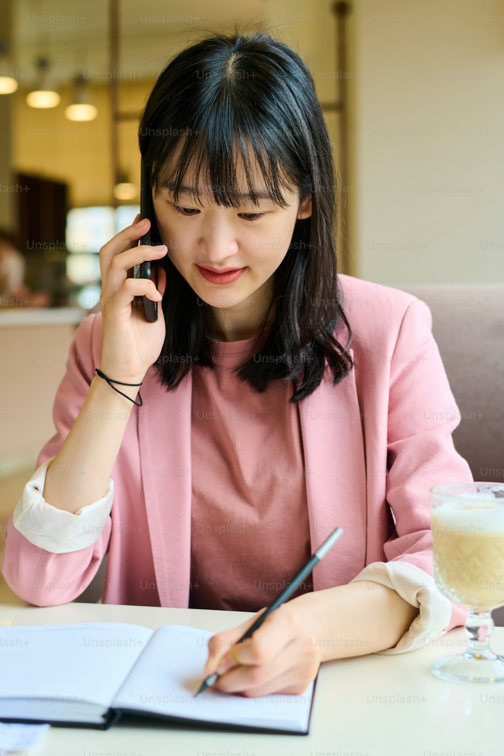 Joven empresaria asiática tomando notas en su agenda durante su conversación en el teléfono móvil en la mesa de la cafetería