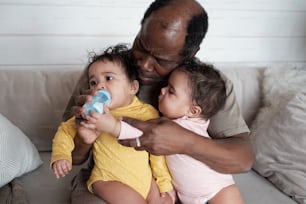 Porträt eines modernen, reifen afroamerikanischen Vaters, der sich um seine Zwillingsbabys kümmert, die eines von ihnen mit der Flasche füttern