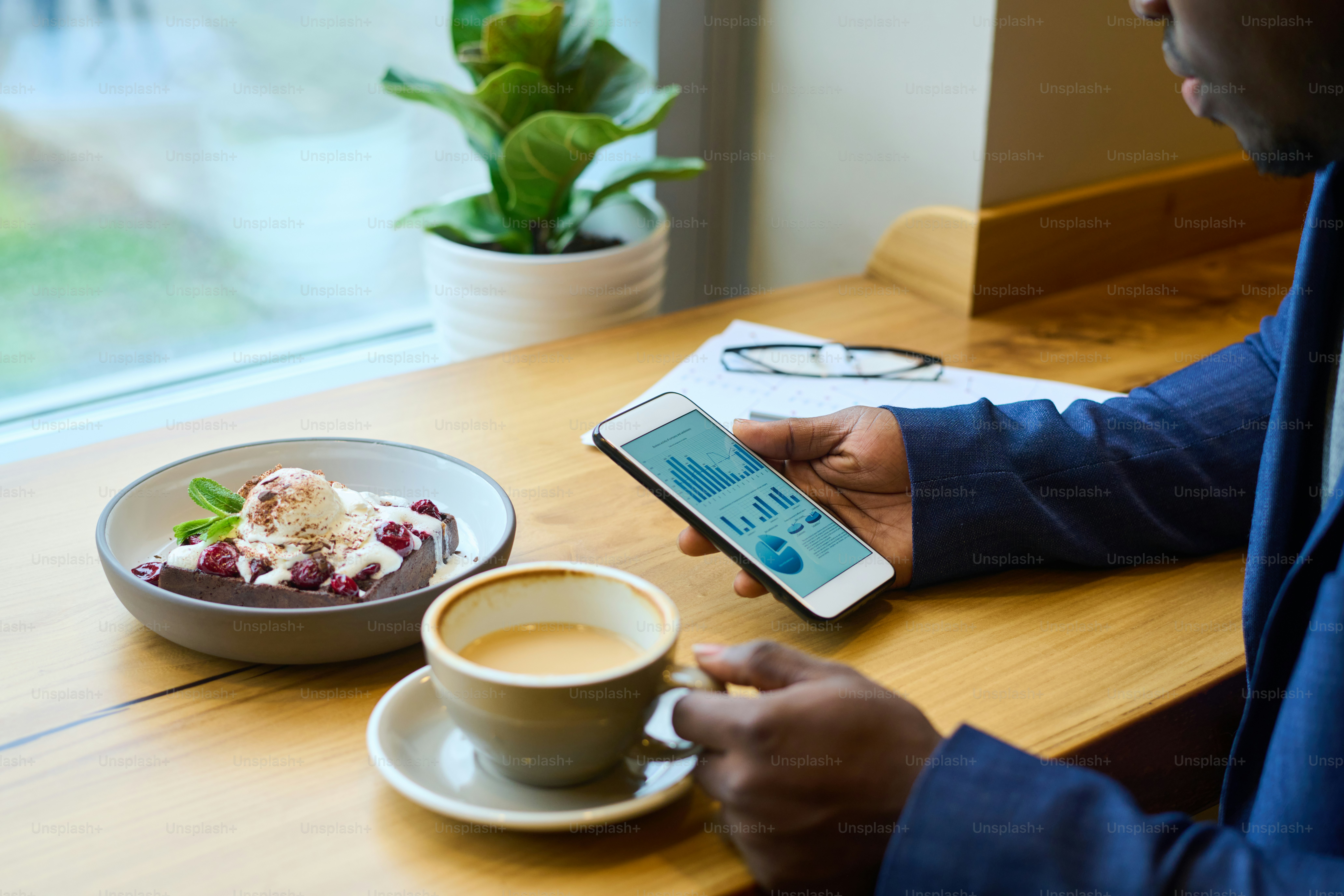 Close-up of African businessman analyzing financial market on his mobile phone while drinking coffee with dessert at table in cafe