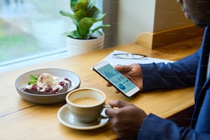 Gros plan d’un homme d’affaires africain analysant le marché financier sur son téléphone portable tout en buvant un café avec un dessert à table dans un café