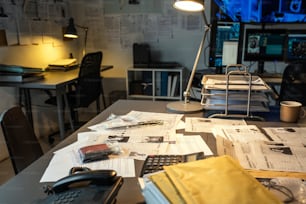 Partie du lieu de travail de l’agent du FBI moderne avec des documents, des preuves, une calculatrice et un téléphone sur un bureau éclairé par une lampe dans un petit bureau