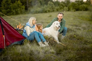 小さな女の子と犬を連れた素敵なカップルの肖像画は、山を旅しながら一緒に座っています。白人の若い家族が自然の中で夏休みを過ごす
