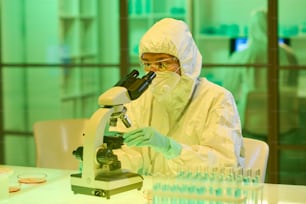Young female scientist in protective biohazard suit, respirator, gloves and eyeglasses looking in microscope while studying new virus or bacteria
