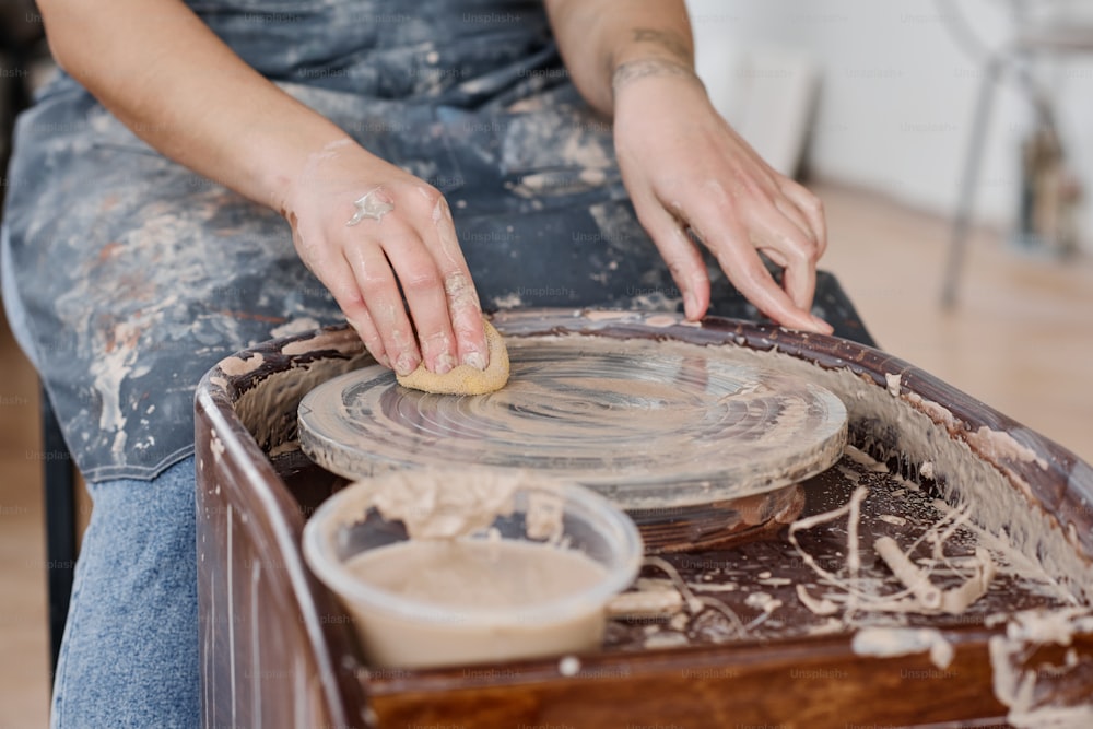 Mãos da mulher nova que limpa a roda de cerâmica com esponja molhada depois de criar novos itens de barro para venda na oficina ou no estúdio