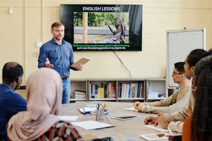 Professor profissional de inglês em pé à mesa na frente de estudantes migrantes multiétnicos explicando-lhes algo