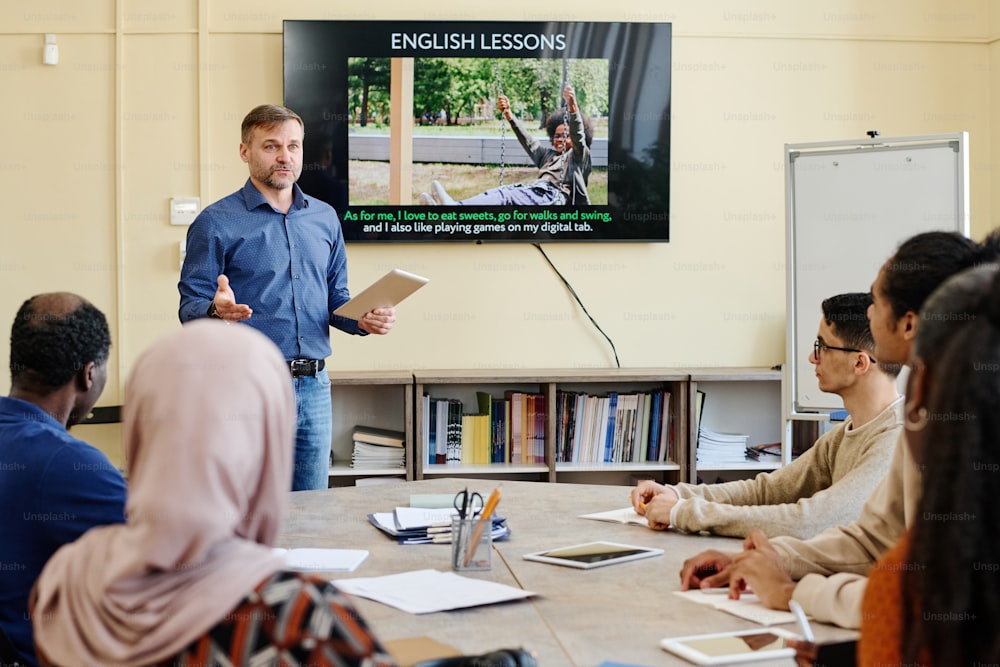 Profesor profesional de inglés parado en la mesa frente a estudiantes migrantes multiétnicos explicándoles algo