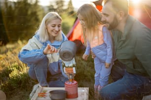 小さな女の子を連れた若いカップルが、パッケージに熱湯を注ぎながら、ハイキング用の乾燥食品を作っています。キャンプ場で旅行者のために料理を作る家族。昇華した食べ物と家族のキャンプのコンセプト