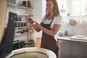 Artesana de pie junto a un horno en su taller de cerámica examinando una pieza de cerámica vidriada recién hecha