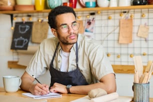 Junger nachdenklicher schwarzer Mann in Arbeitskleidung, der beiseite schaut, während er am Arbeitsplatz sitzt und Skizzen neuer kreativer Artikel im Notizblock zeichnet