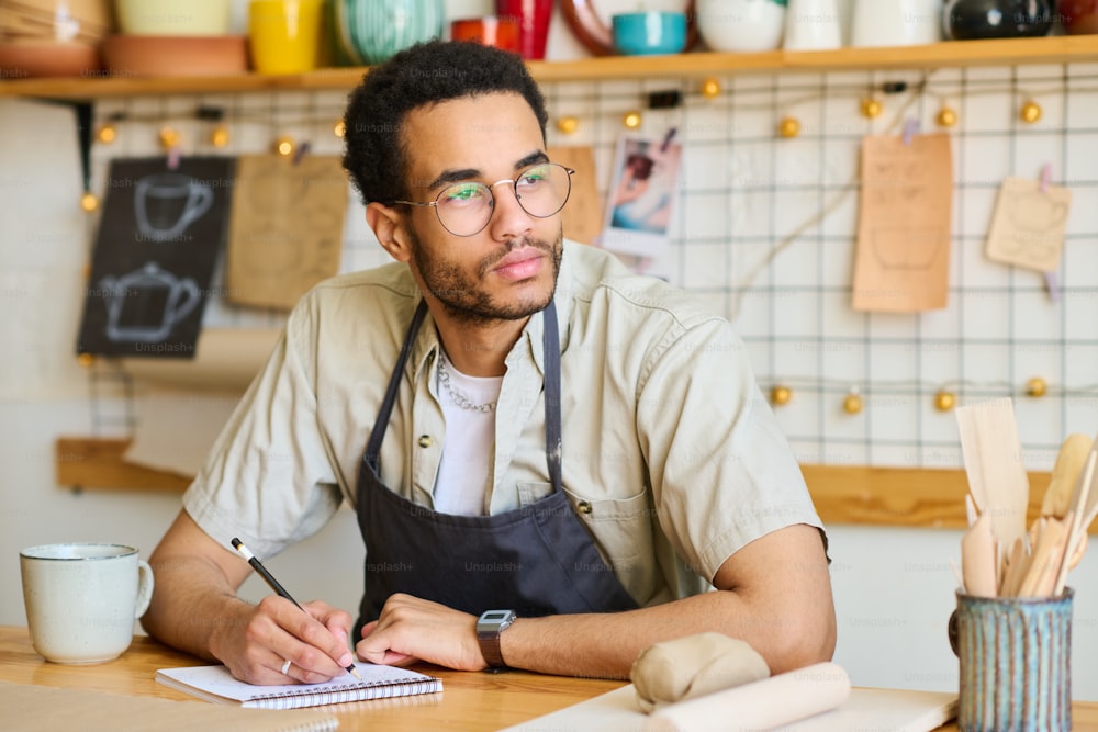 Junger nachdenklicher schwarzer Mann in Arbeitskleidung, der beiseite schaut, während er am Arbeitsplatz sitzt und Skizzen neuer kreativer Artikel im Notizblock zeichnet