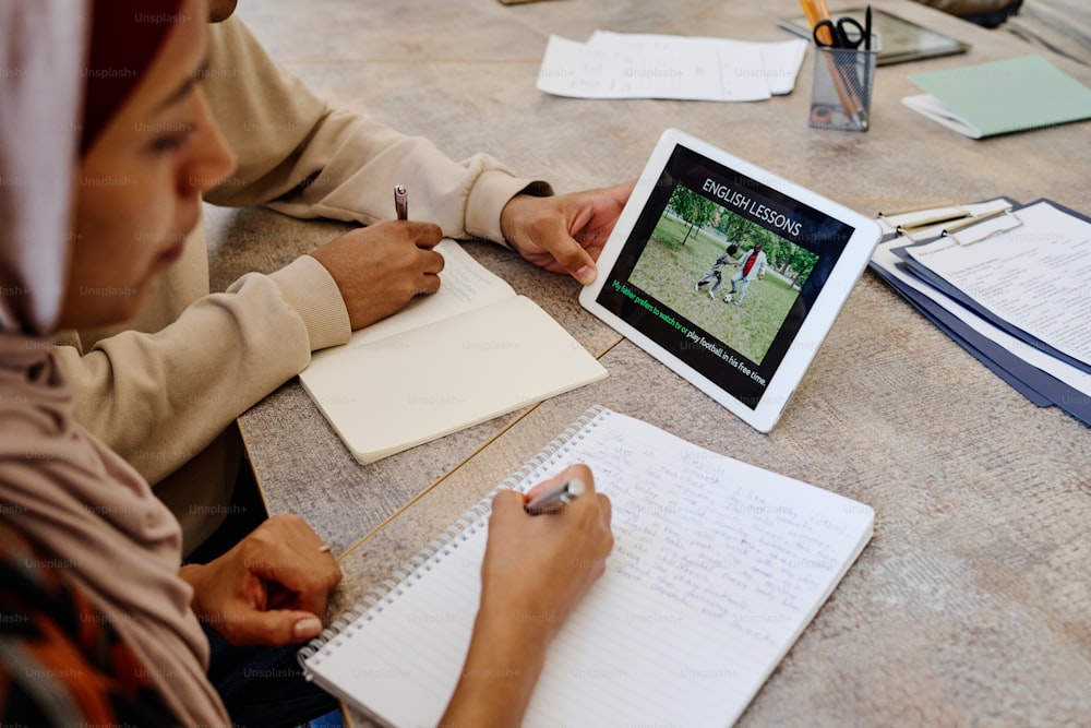 Mulher muçulmana e seu colega de classe irreconhecível assistindo a vídeos de educação em tablet digital e fazendo anotações durante a aula de inglês