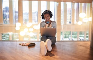 Smiling young African college student sitting on a campus floor reading a text message her cellphone and using a laptop