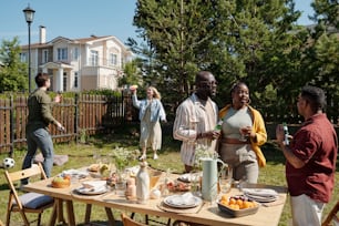 Young black couple with glasses of homemade wine and Hispanic man having chat during picnic against guy and girl playing outdoor game