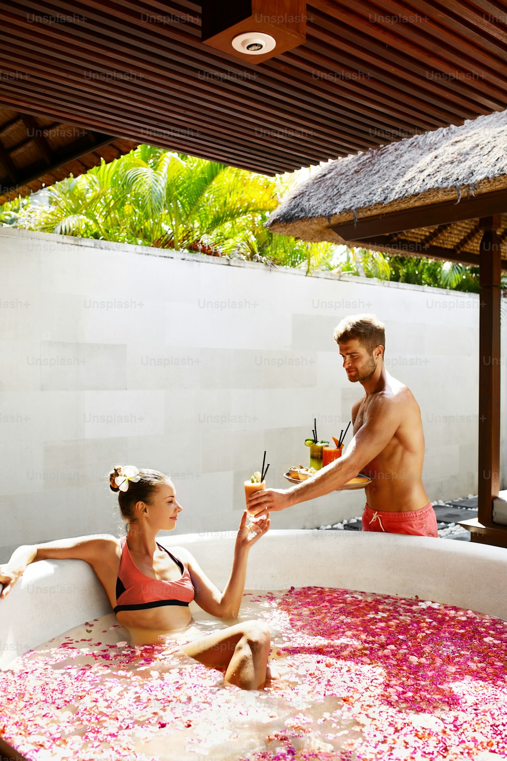 Couple In Love At Spa Resort. Handsome Happy Man Serving Beautiful Smiling Woman Healthy Fresh Detox Juice, Cocktail Drink On Romantic Summer Travel Vacation. Girl Relaxing In Flower Bath At Day Salon