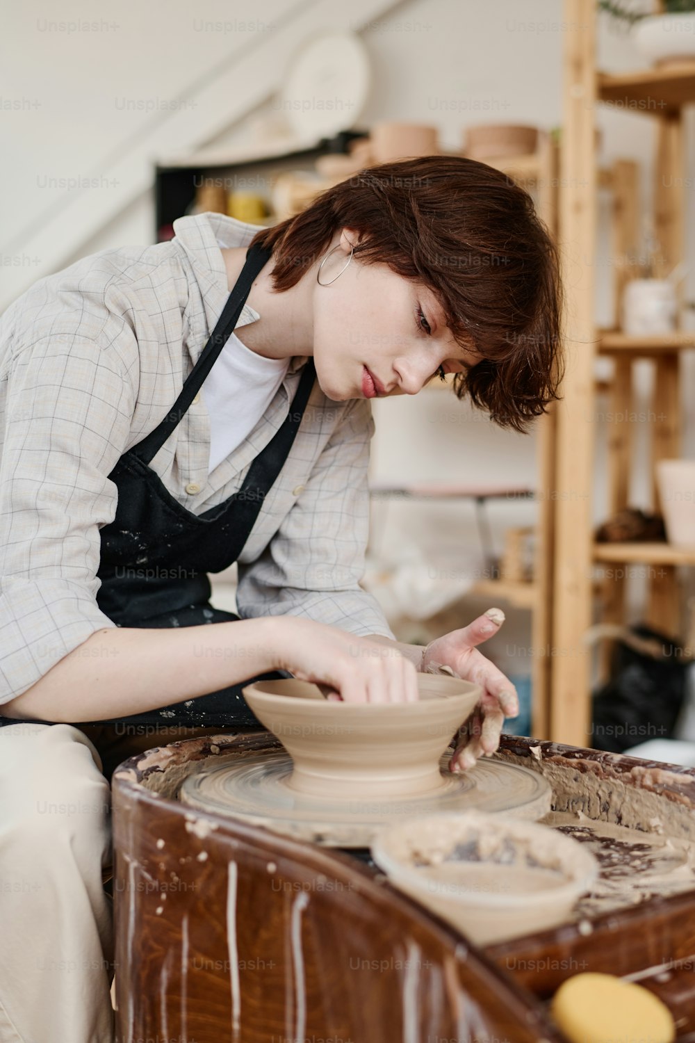 Young female pitcher bending over rotating pottery wheel during work over new clay item while sitting in her workshop or studio