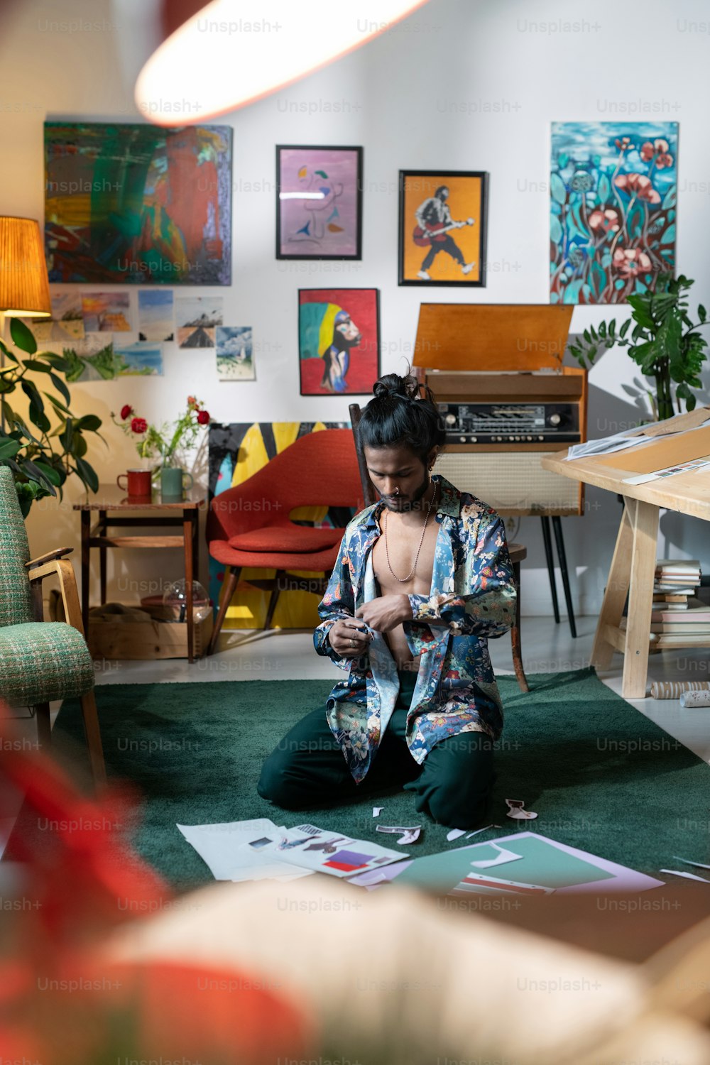 Giovane designer creativo che indossa una camicia sbottonata seduta sul pavimento a casa che lavora su collage ritagliando forme e immagini