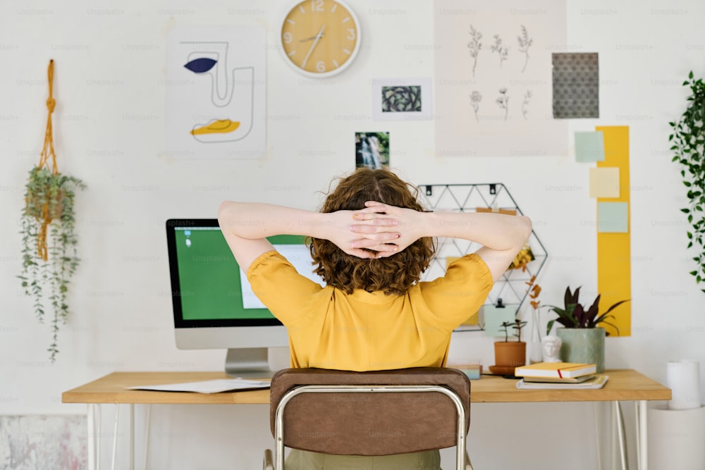 Rückansicht eines jungen freiberuflichen Designers, der eine Minute Ruhe hat, während er vor dem Computermonitor sitzt und die Hände hinter dem Kopf hält