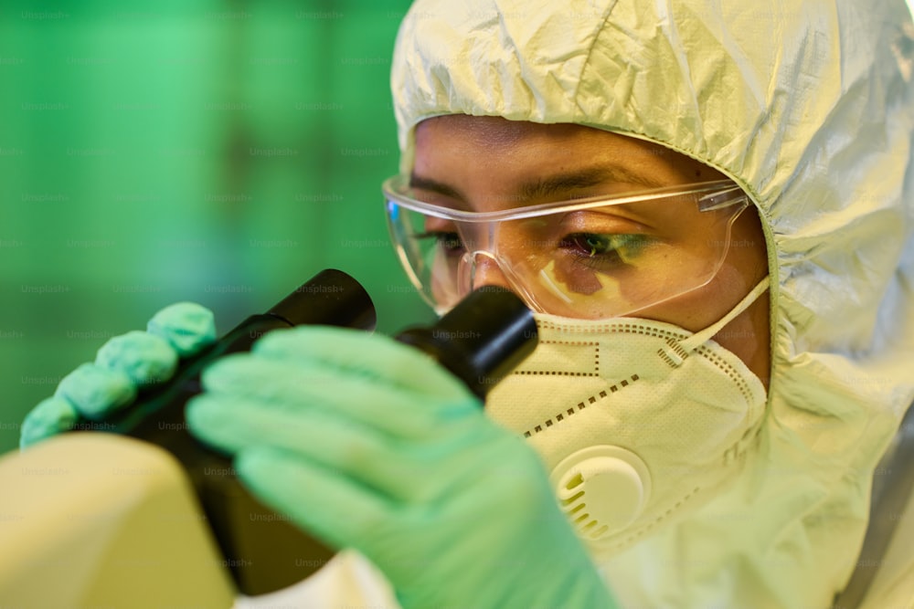 Gloved female virologist in protective coveralls and eyeglasses studying characteristics of new strain of coronavirus in clinical laboratory